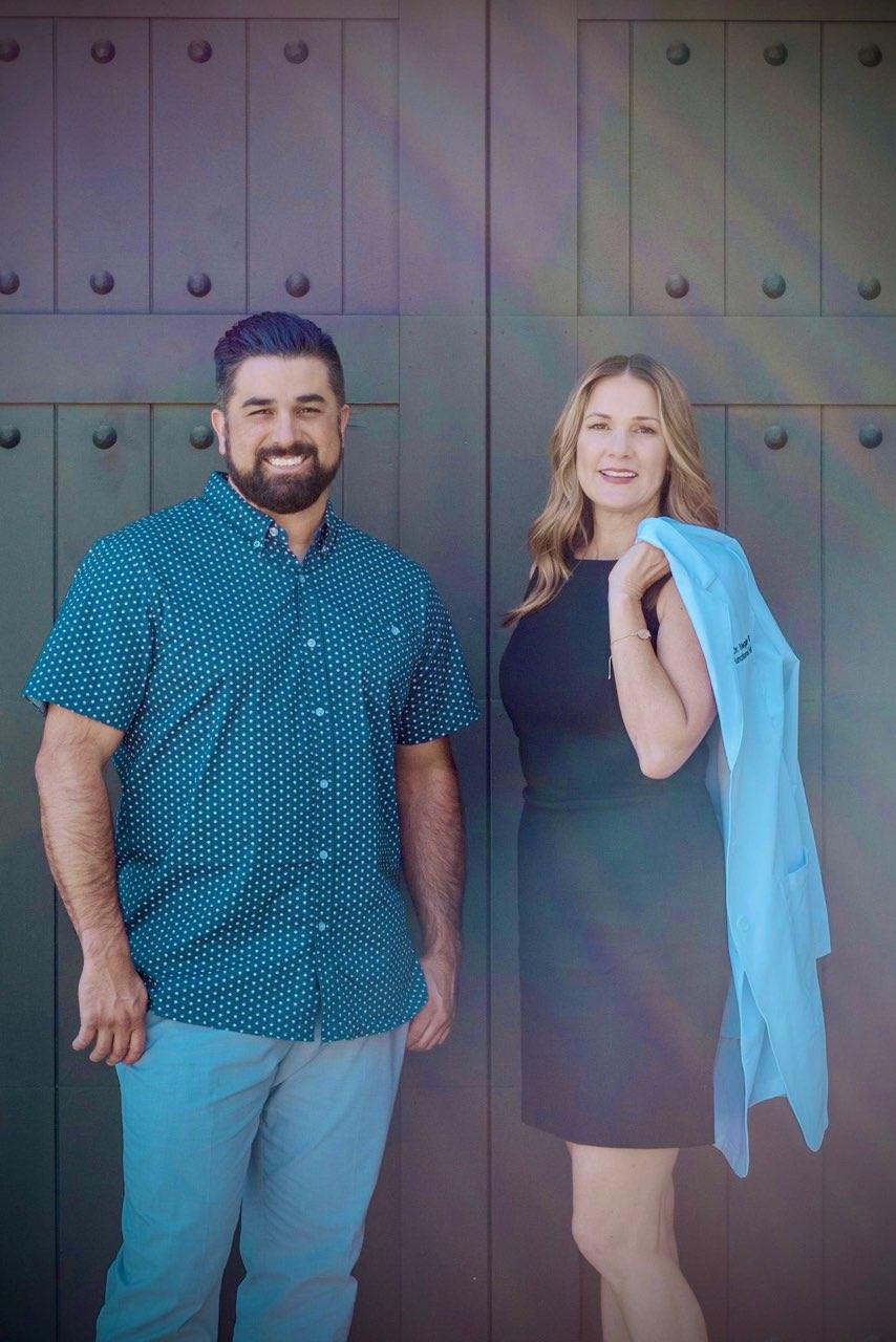 A man in a blue patterned shirt and light pants stands next to a woman in a black dress who is holding a light blue blazer over her shoulder. They are smiling and posing in front of large, dark green doors.