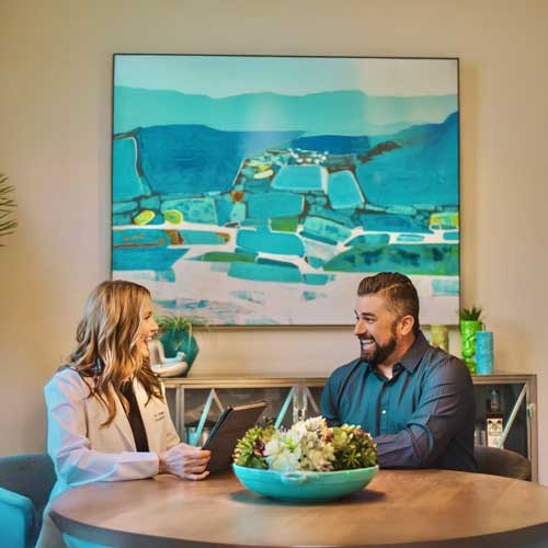 A woman in a white coat and a man sit at a round table, smiling and talking. The woman holds a tablet. Behind them is a large, colorful abstract painting in shades of blue and teal. A bowl of flowers sits on the table.