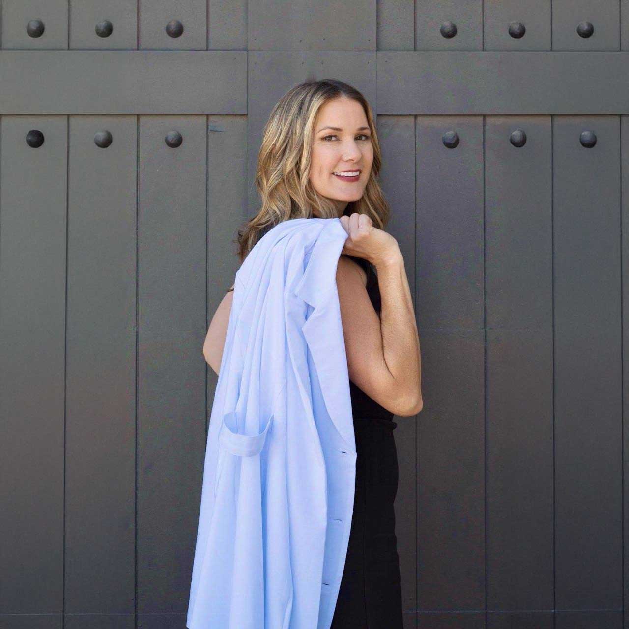 A woman with wavy blonde hair wearing a black outfit stands in front of a large dark door, smiling at the camera while holding a light blue jacket over her shoulder.