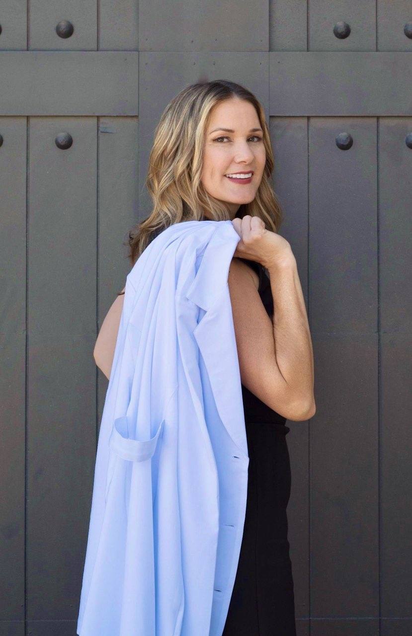 A woman with wavy blonde hair in a black dress smiles at the camera, holding a light blue coat draped over her shoulder, standing in front of a dark wooden door.