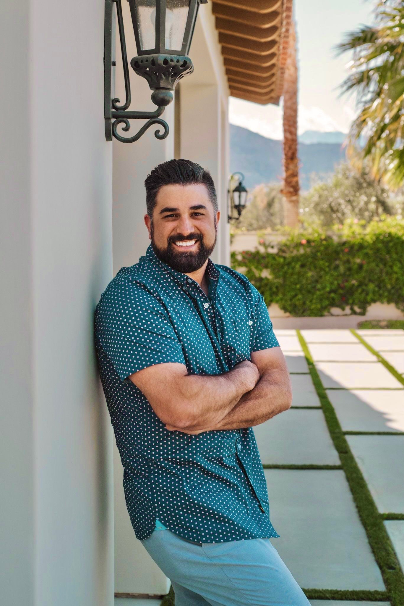A man with dark hair and a beard, wearing a blue short-sleeve shirt with white polka dots, stands smiling with arms crossed against a white wall outdoors, with palm trees and mountains in the background.