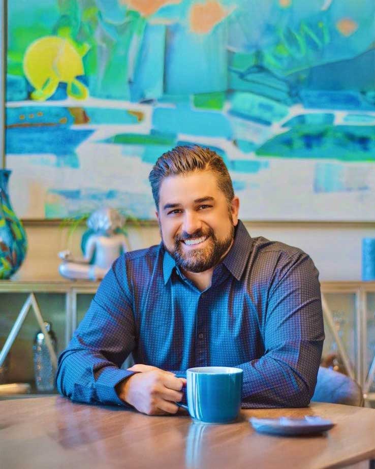 A smiling man with a beard sits at a wooden table holding a blue coffee mug, with colorful abstract artwork and decor in the background. The setting is bright and inviting.