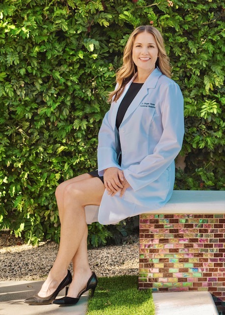A woman wearing a white lab coat and black dress sits on a colorful tiled bench outdoors, smiling, with green leafy plants in the background.