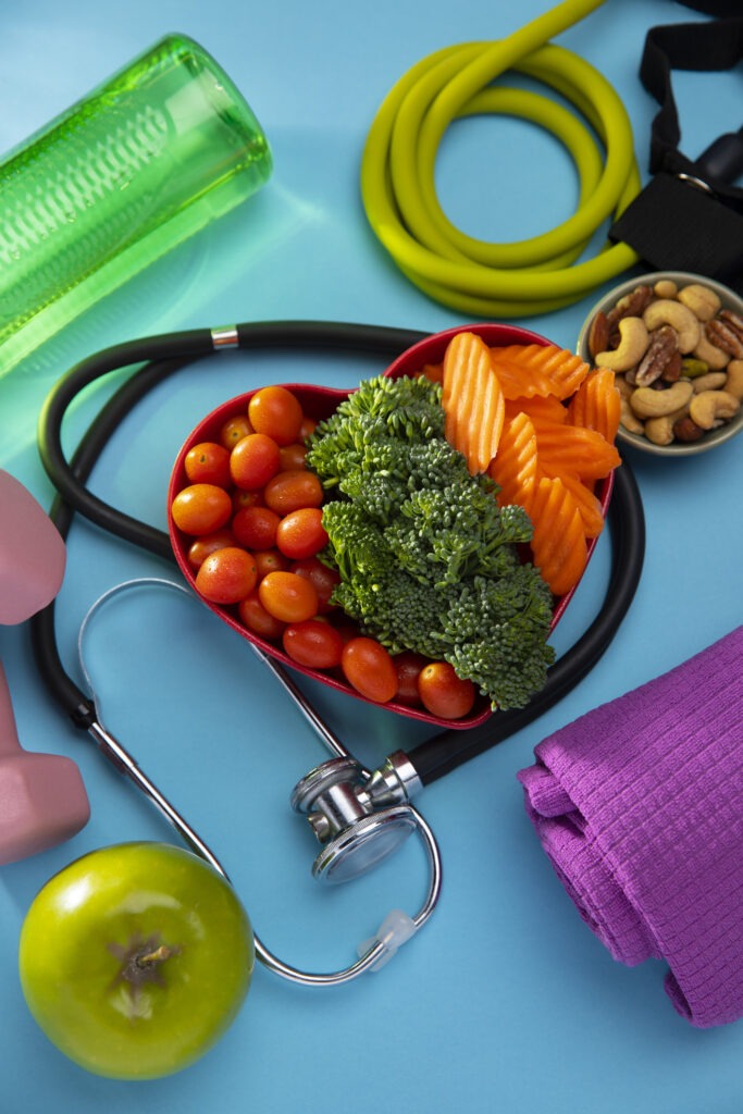 Heart-shaped bowl with tomatoes, broccoli, and carrots surrounded by a stethoscope, pink dumbbells, green apple, yoga mat, green water bottle, jump rope, and a bowl of mixed nuts on a blue background.
