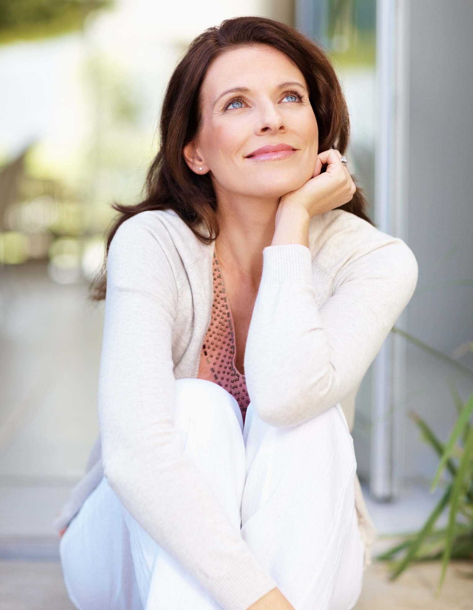 A woman with long brown hair sits outdoors, smiling gently and looking upward. She is wearing a light cardigan and white pants, with one hand resting on her chin. The background is softly blurred.