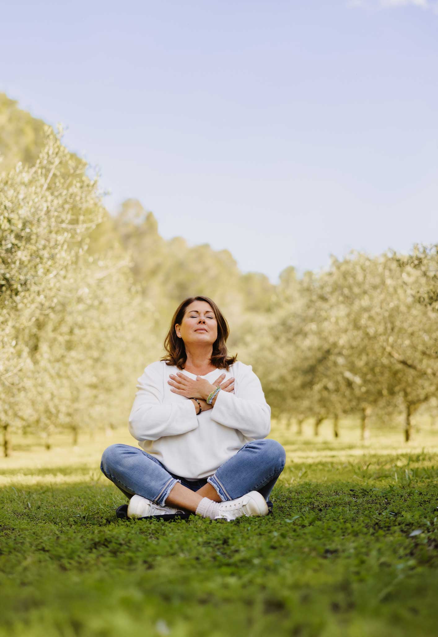 A woman sits cross-legged on grass in an outdoor setting, eyes closed, with her hands crossed over her chest. She appears calm and meditative, surrounded by trees under a clear blue sky.