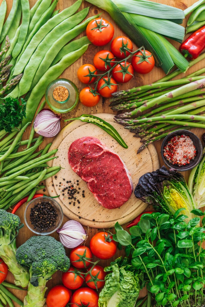 A raw steak on a wooden cutting board surrounded by fresh vegetables including tomatoes, broccoli, asparagus, green beans, lettuce, leeks, red onion, herbs, and bowls of spices and peppercorns.