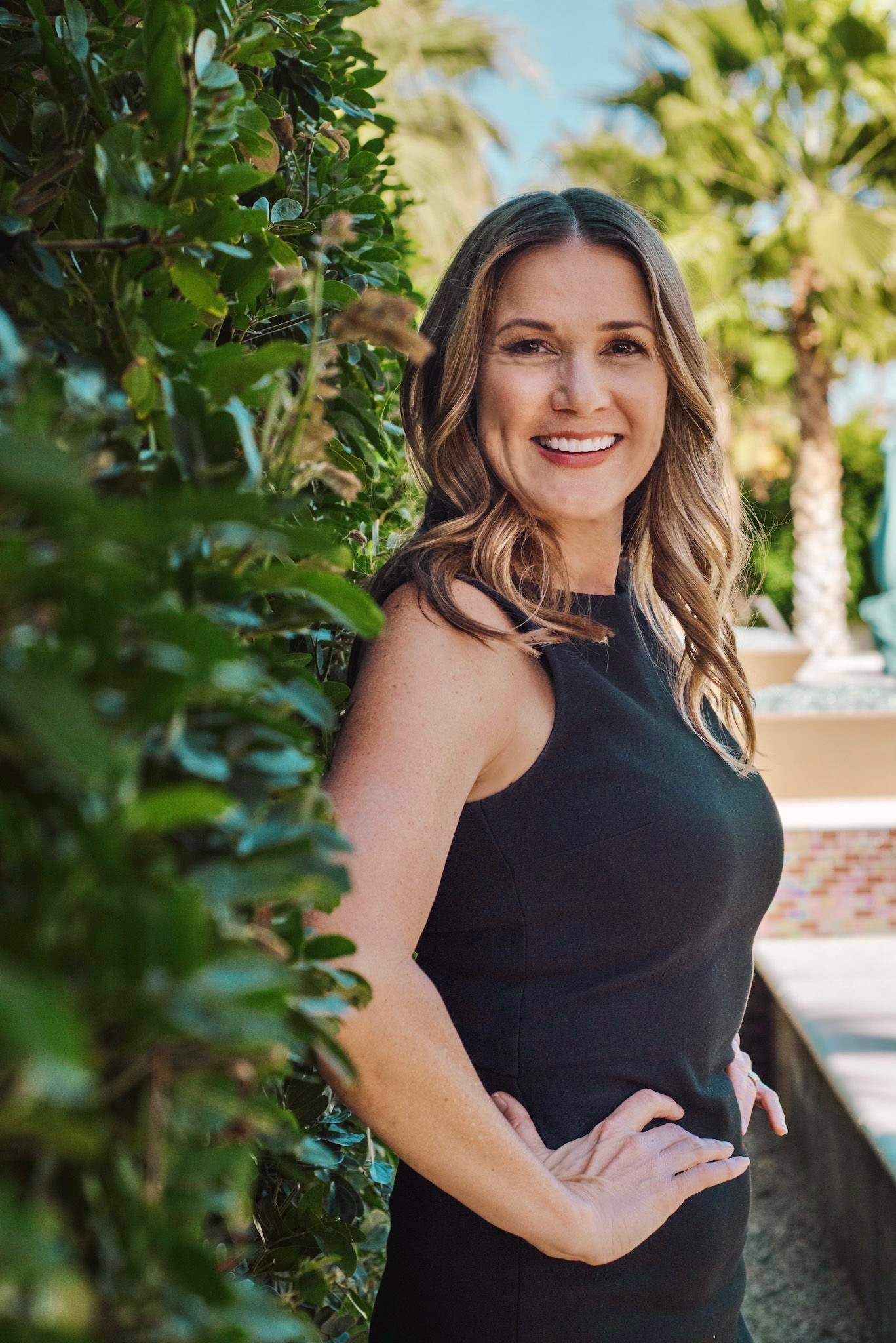 A woman with wavy brown hair, wearing a sleeveless black dress, stands outdoors near green bushes, smiling with one hand on her hip. Palm trees and a sunny sky are visible in the background.