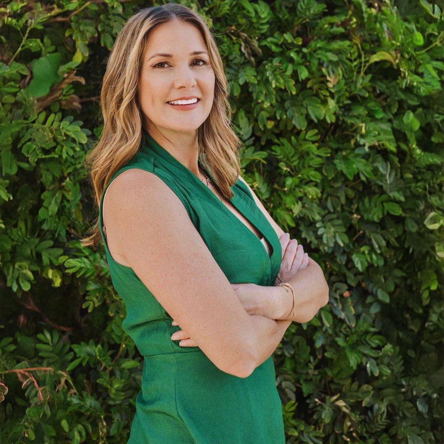 A woman with wavy light brown hair, wearing a sleeveless green dress, stands outdoors with her arms crossed and smiles at the camera. Lush green foliage fills the background.