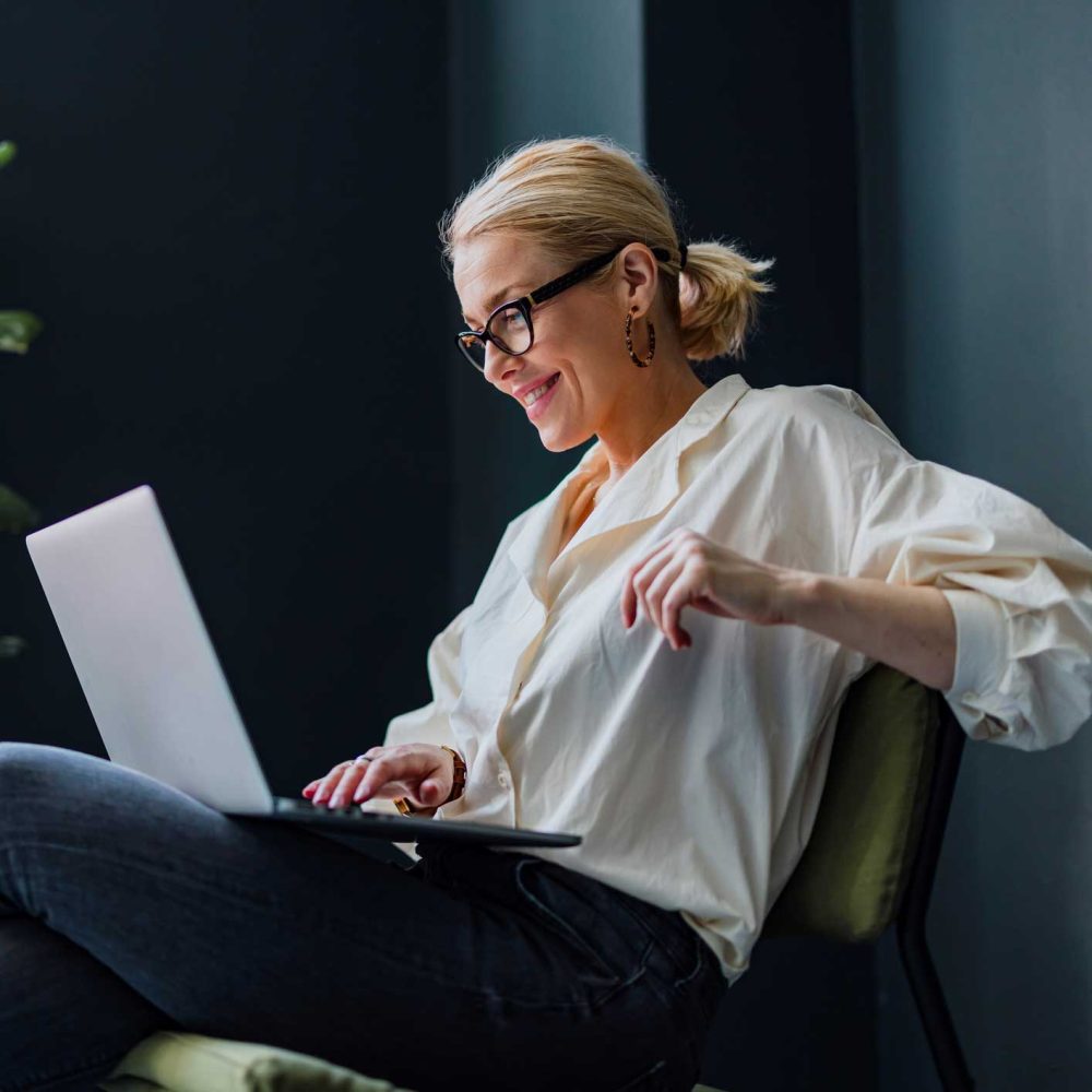 A smiling woman with glasses and a white shirt sits on a chair using a laptop, appearing relaxed and engaged in her work.