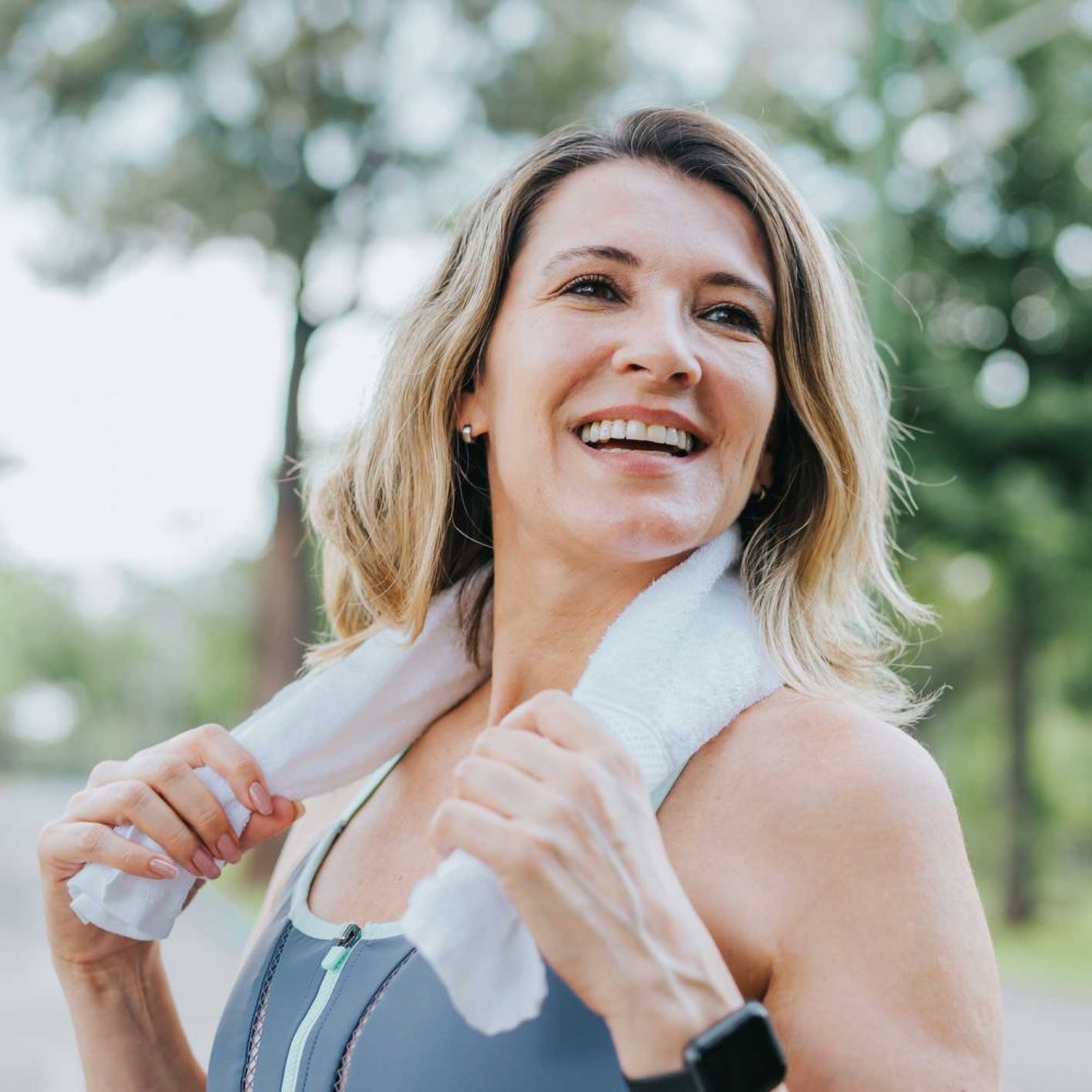 A smiling woman outdoors in athletic wear holds a white towel around her neck. She appears happy and relaxed, with trees and greenery in the blurred background.